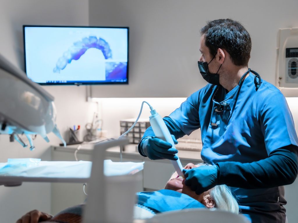 Dentist in blue scrubs using an intraoral scanner, with a 3D scan of the patient's teeth on a wall monitor